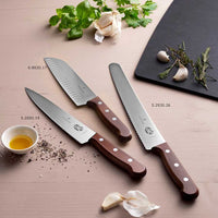 Victorinox Santoku kokkiveitsi ovaalihiottu puu 17 cm osana veitsisarjaa, esillä yrttien ja valkosipulin kanssa keittiössä.