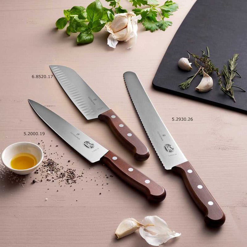 Victorinox Santoku kokkiveitsi ovaalihiottu puu 17 cm osana veitsisarjaa, esillä yrttien ja valkosipulin kanssa keittiössä.