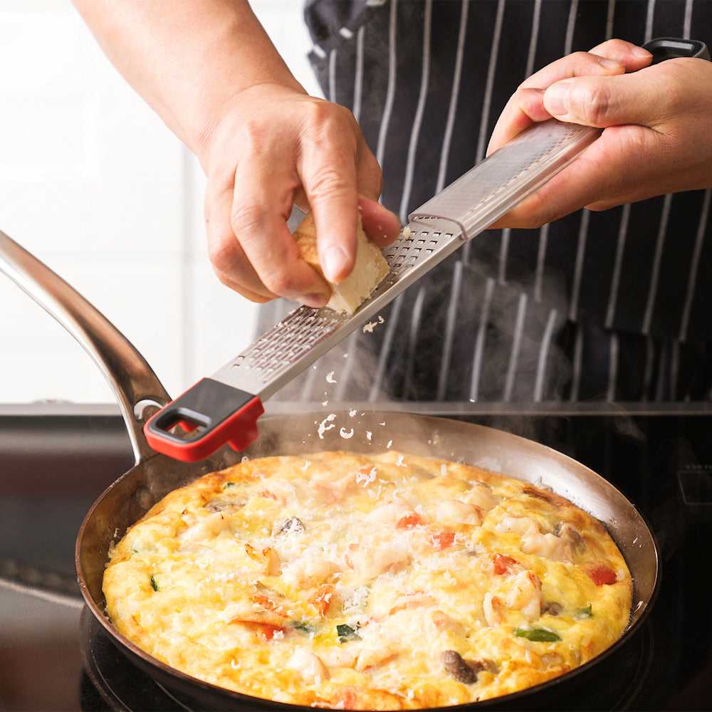 Cuisipro SGT Deluxe Dual Raastin käytössä juuston raastamiseen kuuman munakkaan päälle, monipuolinen keittiöväline ruoanlaittoon.