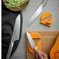 Global 40 v. juhlasetin veitsillä leikataan kurpitsaa – kolme veistä ja leikkuulauta, oranssi kurpitsa ja vihreä salaatti.