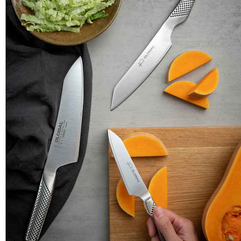Global 40 v. juhlasetin veitsillä leikataan kurpitsaa – kolme veistä ja leikkuulauta, oranssi kurpitsa ja vihreä salaatti.