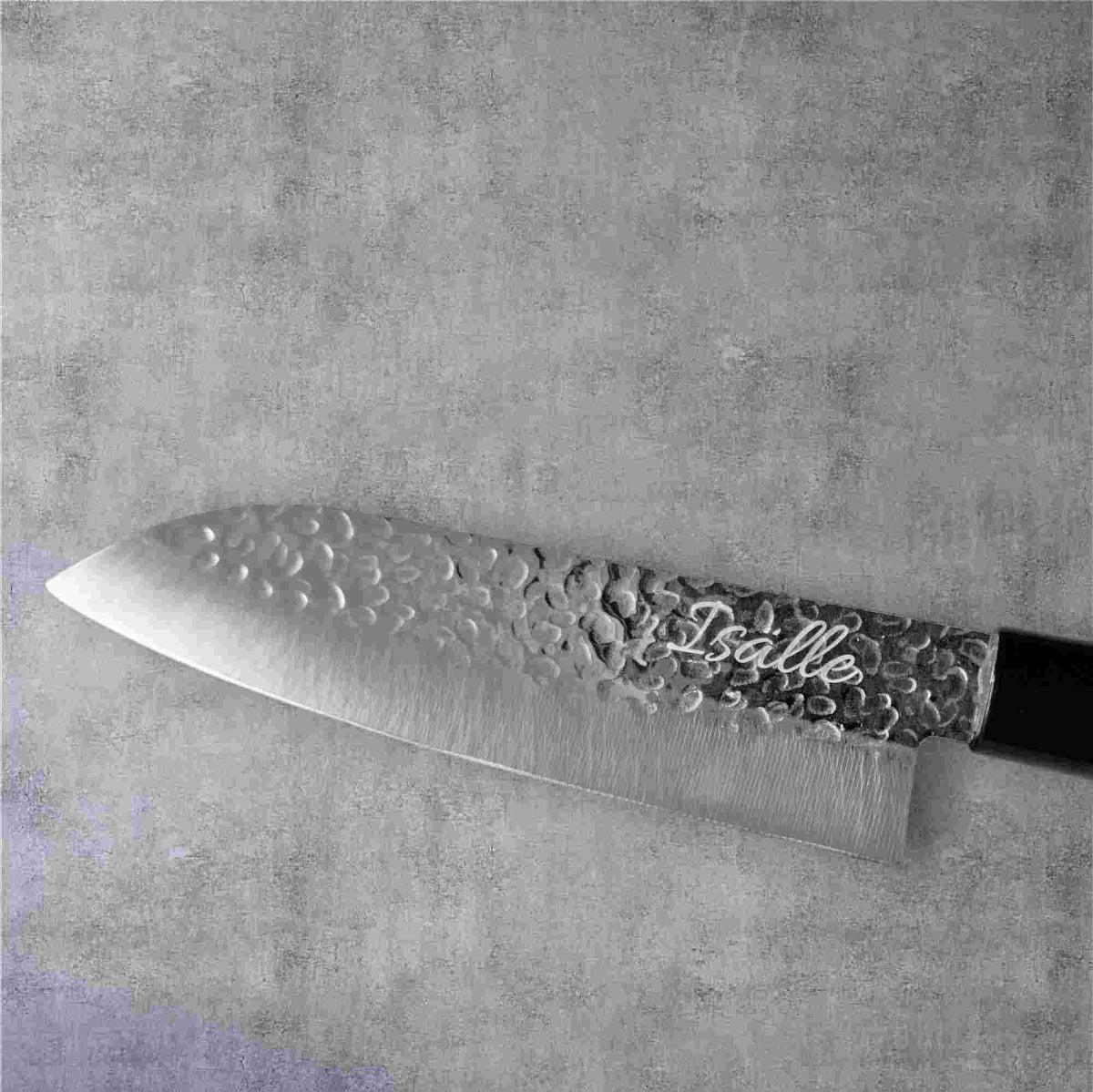 Japanilainen Santoku-veitsi, jossa on vasaroitu terä ja kaiverrettuIsalle"-teksti, harmaalla kivimäisellä taustalla.
