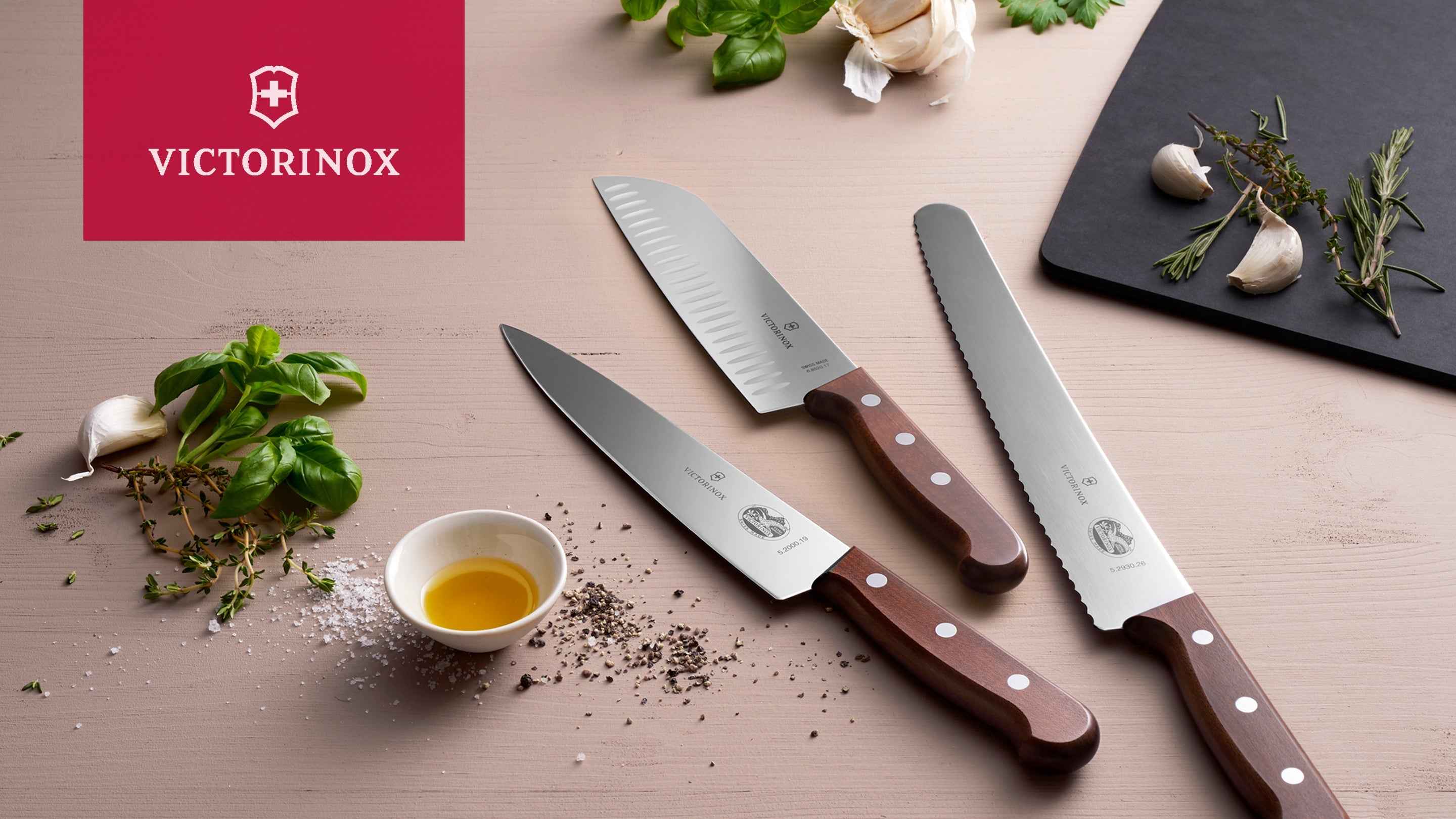 Victorinox-keittiöveitset puisilla kahvoilla, aseteltuna mausteiden, yrttien ja valkosipulin kanssa vaalealle puupöydälle.