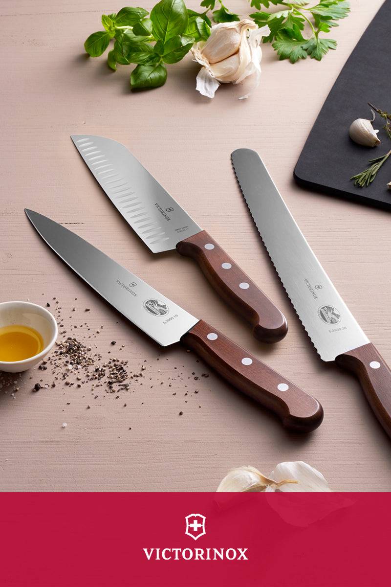 Kolme Victorinox-keittiöveistä puisilla kahvoilla puupöydällä, ympärillä valkosipulia, yrttejä, öljyä ja mustapippuria.