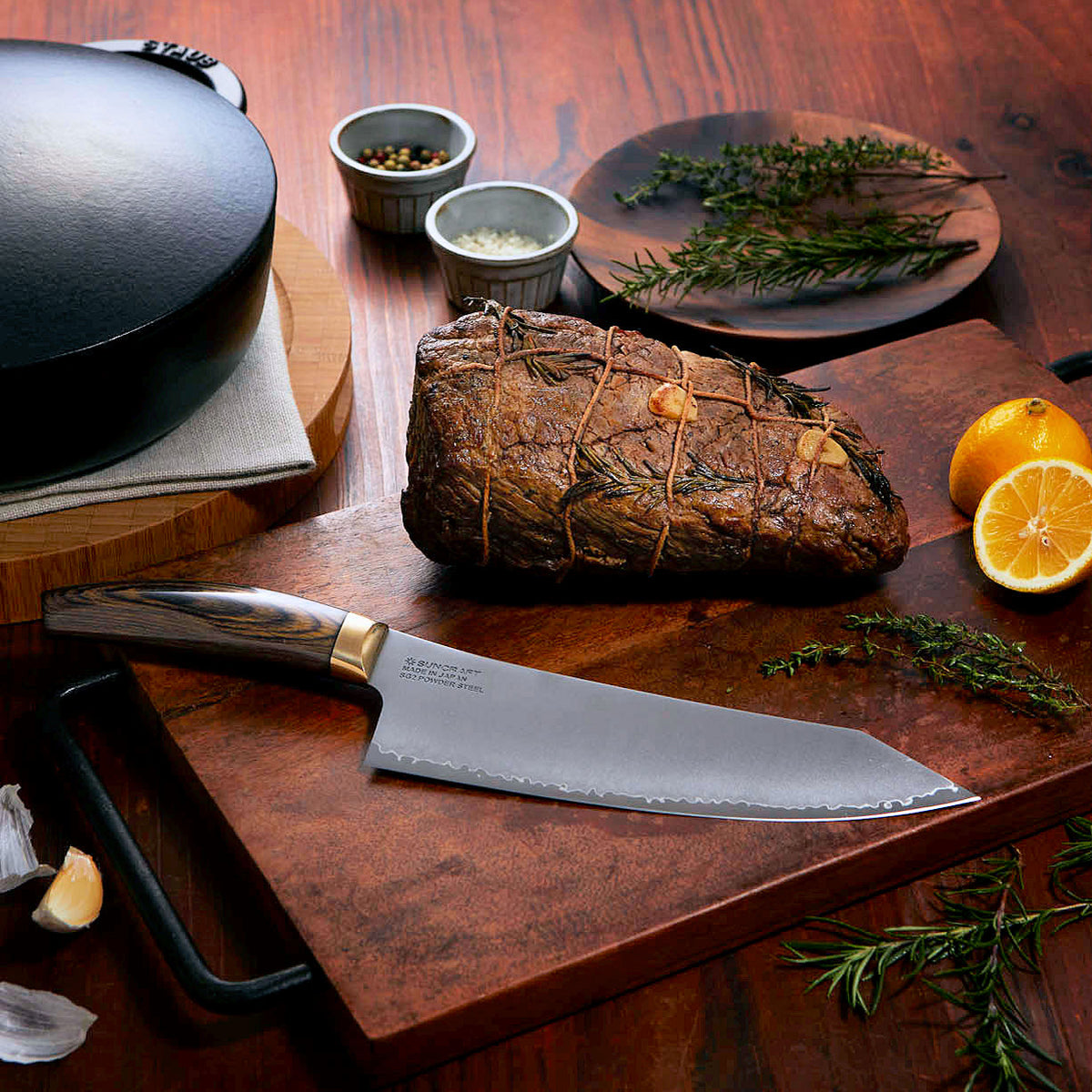 Suncraft Elegancia kokkiveitsi leikkuulaudalla, ympärillä yrttejä, sitruuna ja valkosipulia. SG2-teräs ja Damascus-pinta.