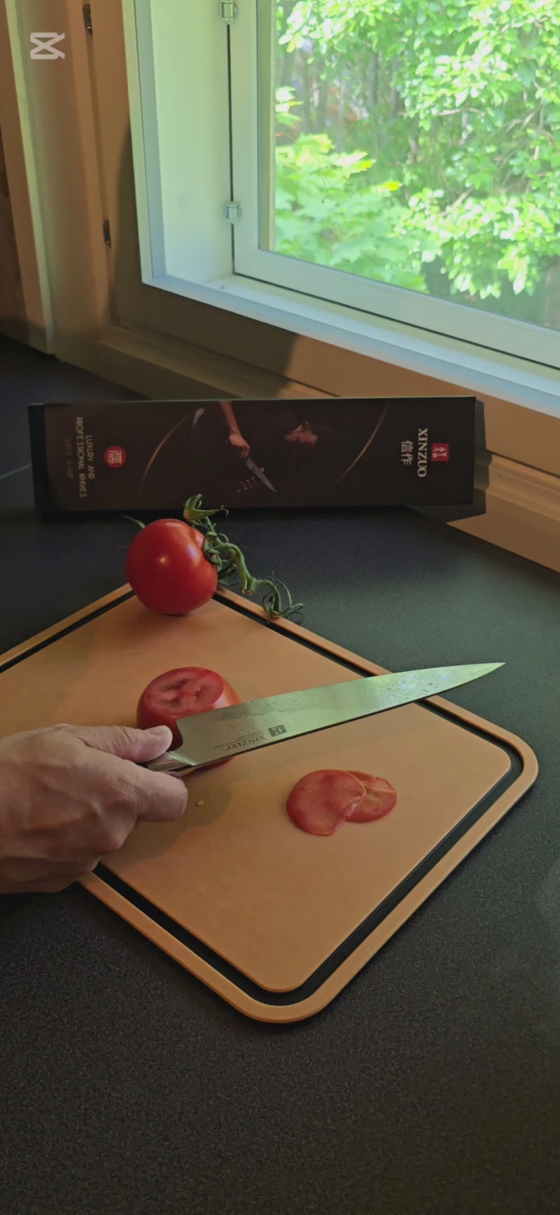 XINZUO Jiang Damaskus Santokuveitsi, 18 cm, viipaloi tomaattia leikkuulaudalla luonnonvalossa, osa keittiövarusteita.